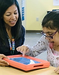 Mai Ling is sitting with a student and holding an AAC device while she access is with her pointer finger.
