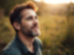 Smiling bearded man gazes upward in a sunlit field at sunset, with warm golden blur behind him.