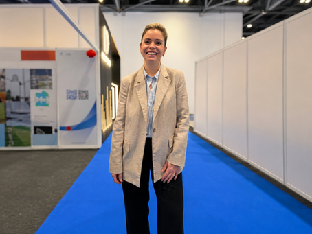 Photo of Carly Taylor, Director of Growth and Customer Experience at IDGateway, smiling at a trade show or exhibition, standing on a blue carpeted aisle with exhibitor stands in the background.
