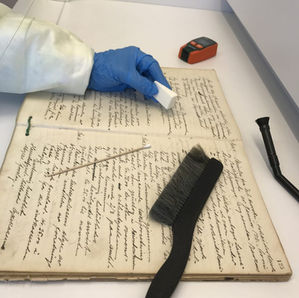 Nærbilde av en hånd med blå plasthanske som holder en hvit liten svamp. Hånden hviler på en oppslått protokoll med gammel håndskrift. På den oppslåtte siden ligger en kost og en pinne med bomullsdott i enden. Ved siden av protokollen ligger endestykket på en liten støvsuger og e luftmåler.
