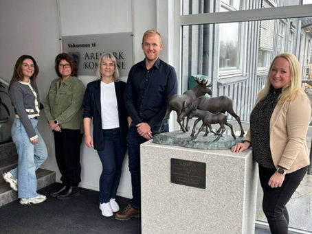 Til venstre i bildet står tre kvinner og en mann stilt opp ved siden av hverandre. Mellom dem og en kvinne til høyre i bildet står en sokkel med statue av tre elger. 