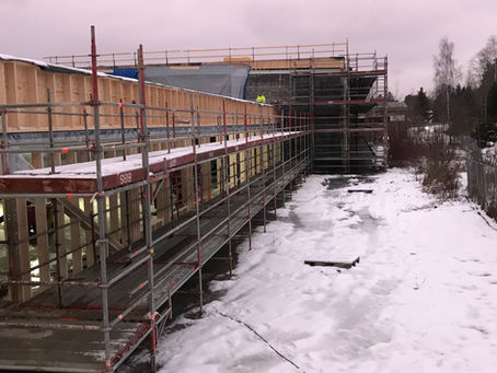 Bilde av en stor bygning fra utsiden. Bygget er under bygging med stillaser rundt hele. På bakken er det snø. En mann med gul jakke står på taket til bygningen.