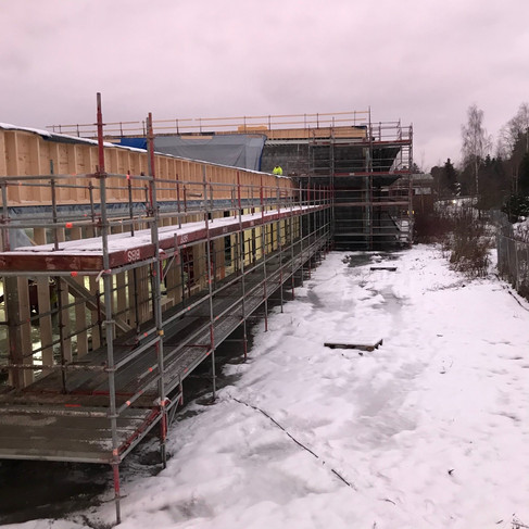 Bilde av en stor bygning fra utsiden. Bygget er under bygging med stillaser rundt hele. På bakken er det snø. En mann med gul jakke står på taket til bygningen.