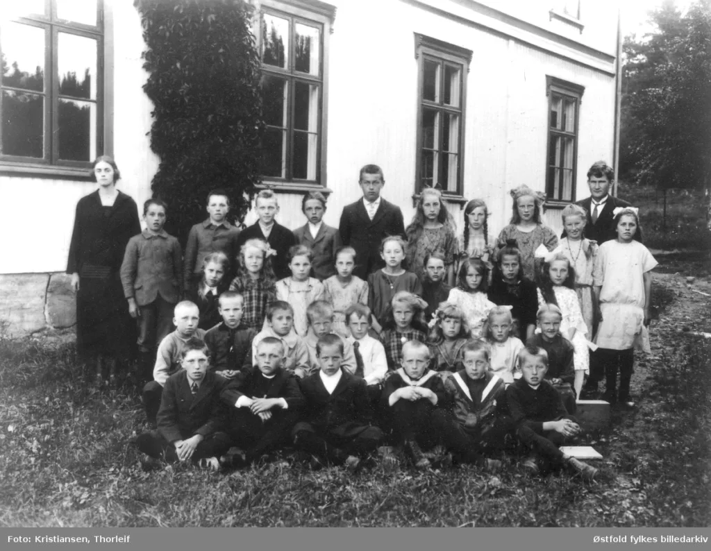Svart/hvit fotografi av en skoleklasse. Guttene er kledd i svart, og jentene har kjole. Til venstre i bildet står en voksen kvinne og til høyre står en voksen mann. Gruppen med elever er plasser foran skolebygget.