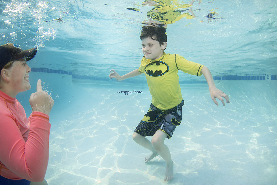 Underwater photography image of a swim instructor showing a child in a batman swimsuit how to smile underwater