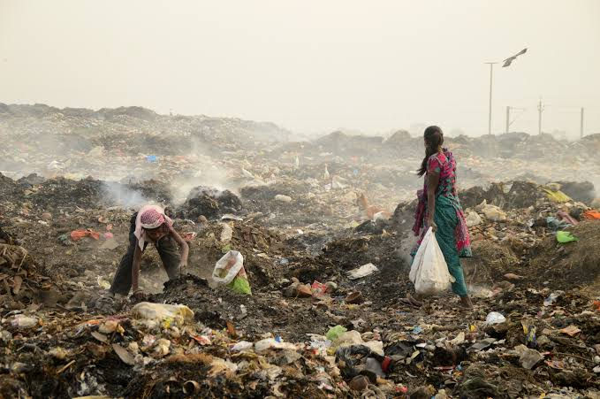 Garbage Collectors searching through a vast garbage dump under a hazy sky, holding bags. Smoke rises from the waste, creating a somber atmosphere.
