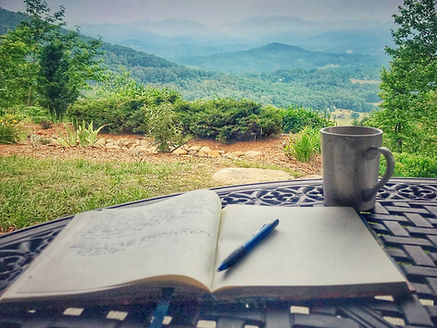 An open journal, pen and cup sitting on a table overlooking mountain vista.