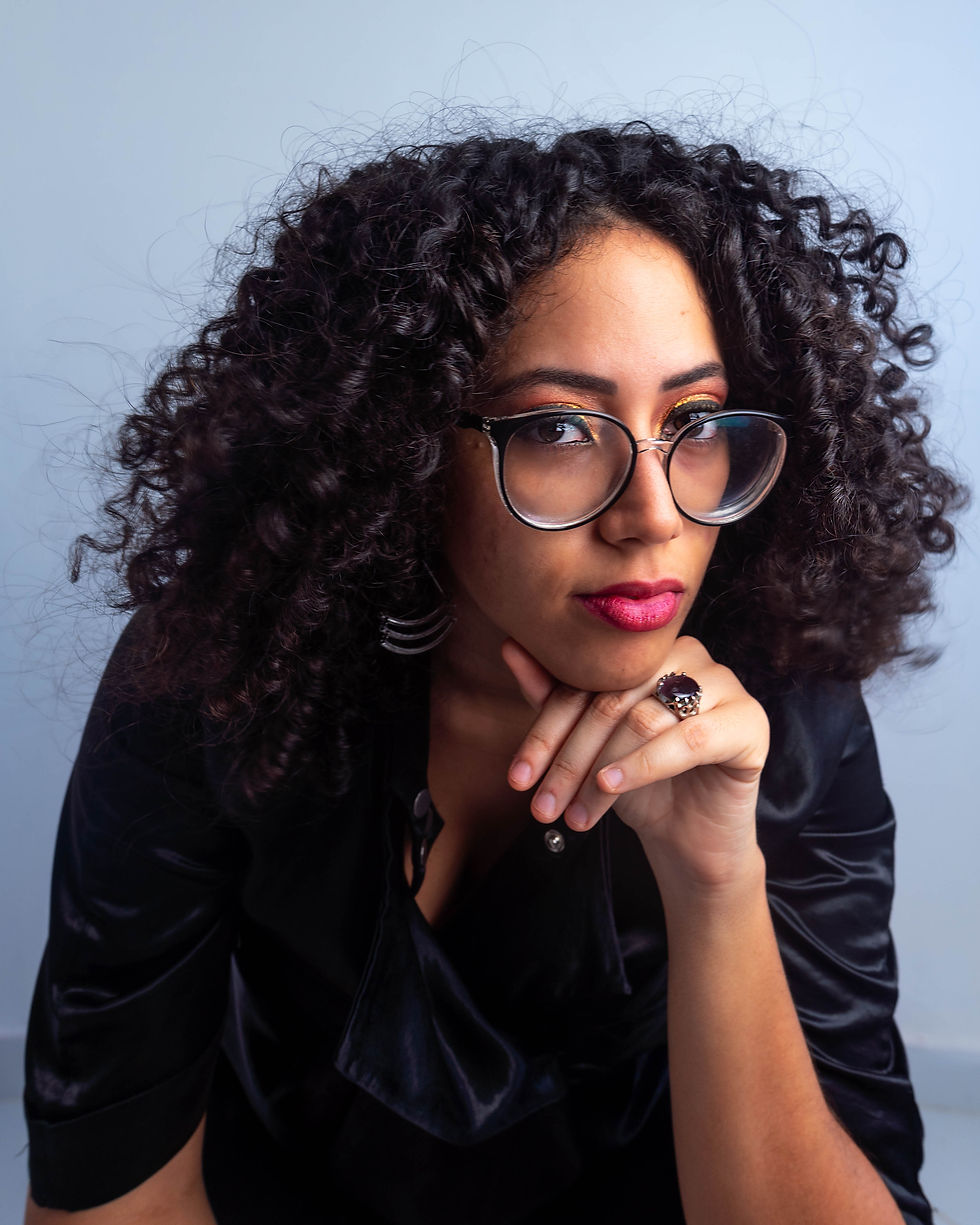 Lara Miranda. Mulher parda, de cabelos cacheados. Usa óculos e roupas pretas enquanto olha séria.