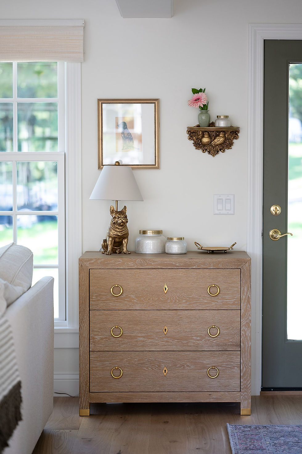 Wooden dresser with gold handles, adorned with a dog statue, jars, lamp. Shelf above holds pink flower in vase. Window and door nearby.