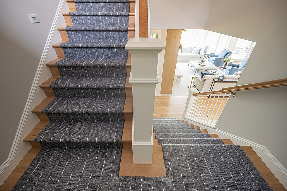 Carpeted staircase with gray runner descends to a bright living room with blue and white chairs and a sofa in a modern home.