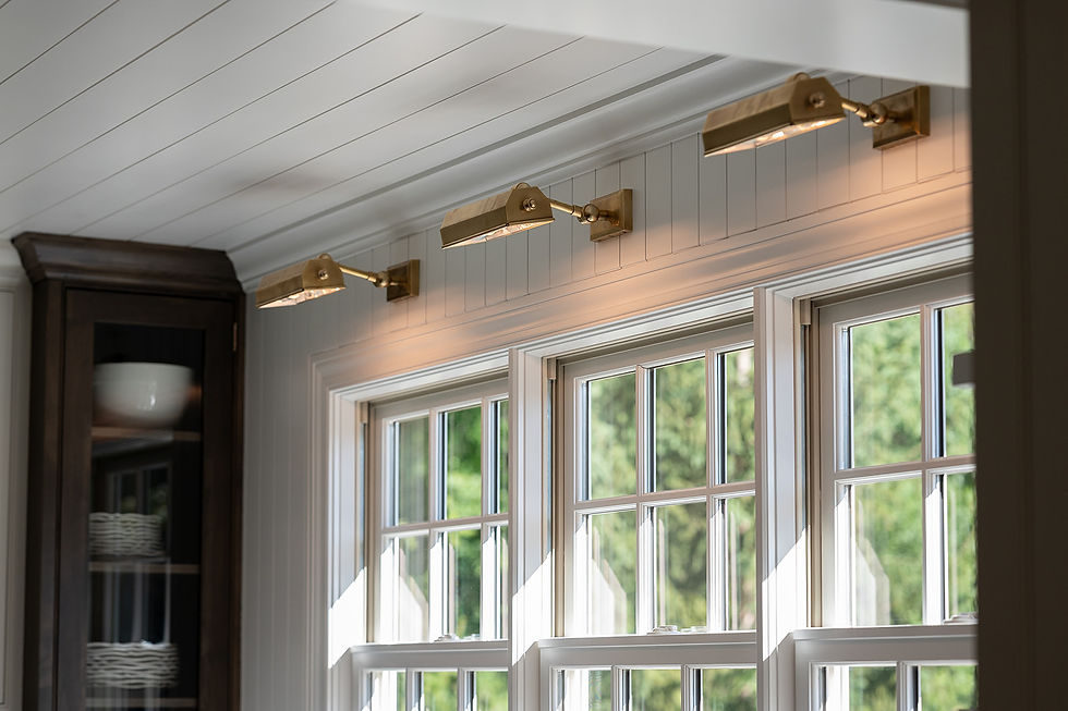 Brass wall lamps above large windows in a bright room with white paneled walls. A cabinet is visible on the left. Natural light filters in.