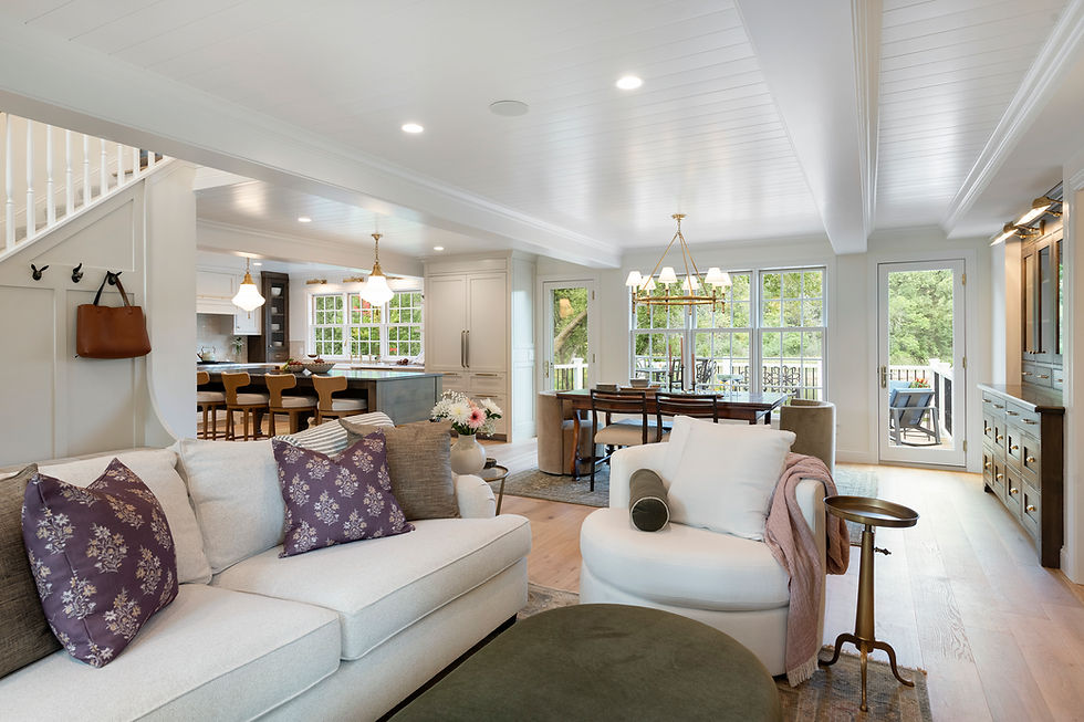 Spacious living room with white walls, cozy sofas with purple pillows, wooden dining table, and large windows overlooking greenery. Bright and inviting.