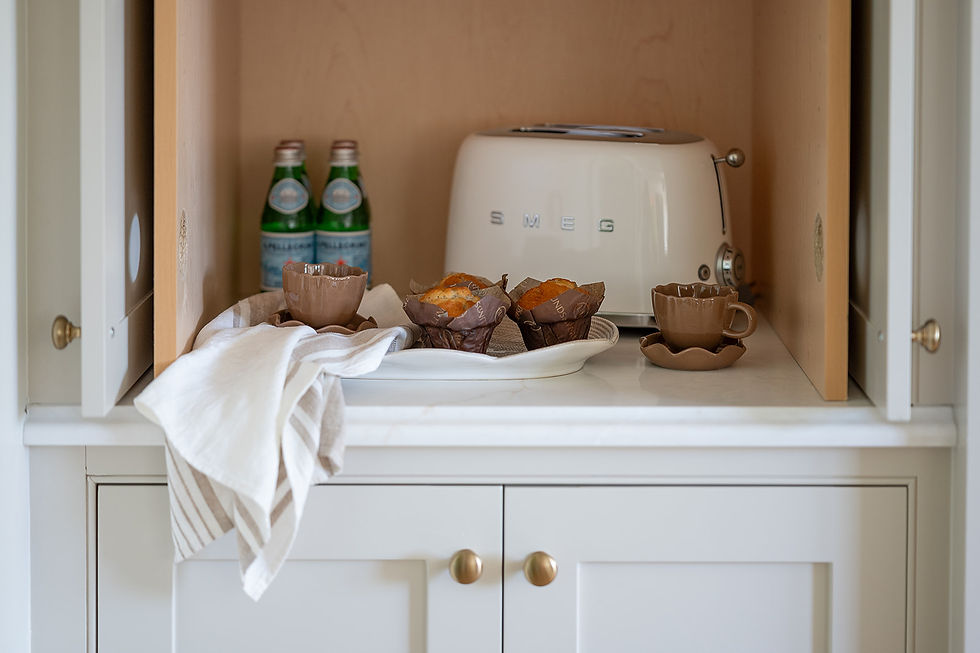 Cups and muffins on a tray beside a white SMEG toaster in a kitchen cupboard. Two bottles in the background, cloth draped in front. Cozy setting.
