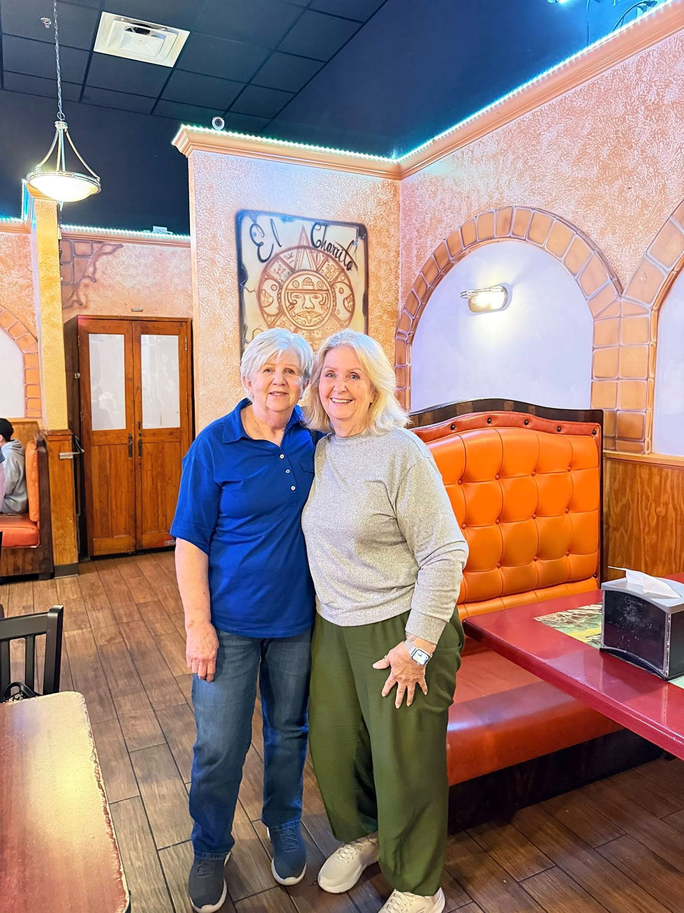 Two smiling women stand close in a restaurant with orange booths. Sign reads "El Charrito" on a decorated wall. Warm, inviting atmosphere.