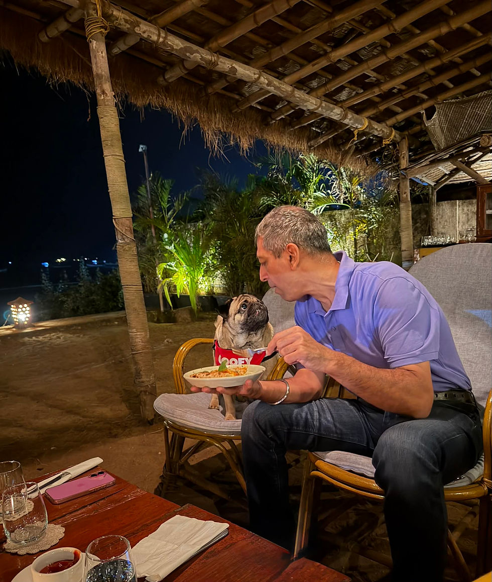 Man in a purple shirt shares food with a pug wearing a "GOOFY" shirt at an outdoor table under a thatched roof, creating a warm ambiance.