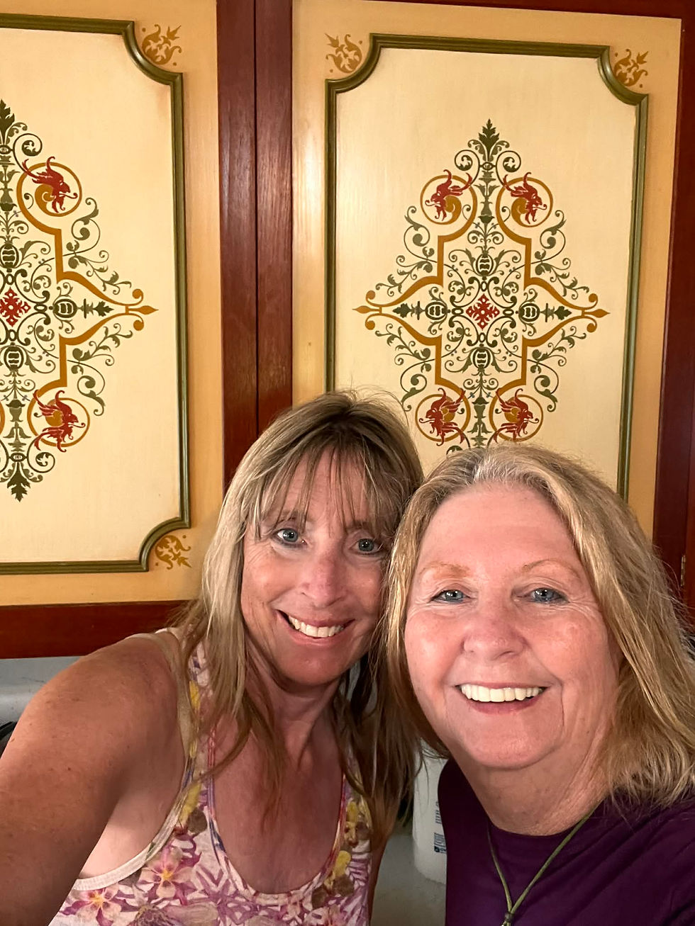 Two smiling women take a selfie in front of ornate cabinet doors with intricate red and green floral designs, creating a warm and cheerful mood.