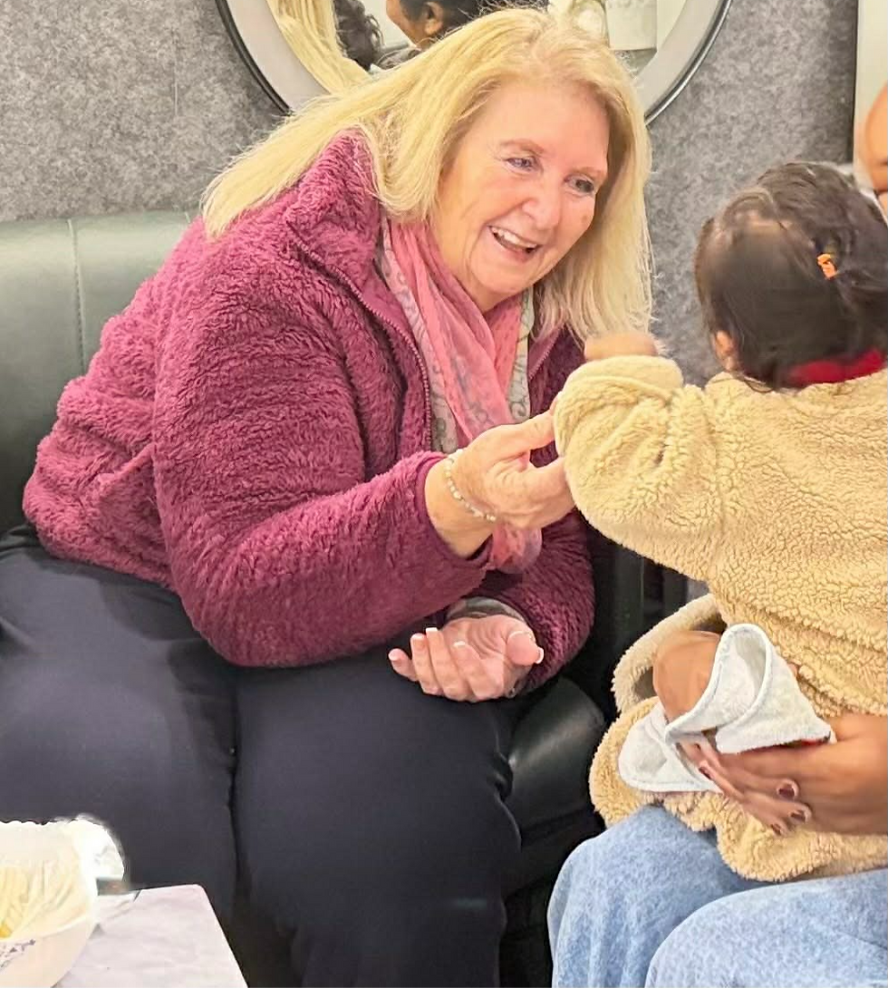 Woman in a maroon jacket smiles at a child in a beige coat. They hold hands in a cozy setting. Mirror and gray wall in background.