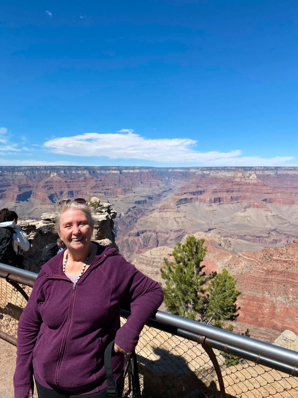 Smiling woman in a purple hoodie posing at a Grand Canyon overlook under a bright blue sky.