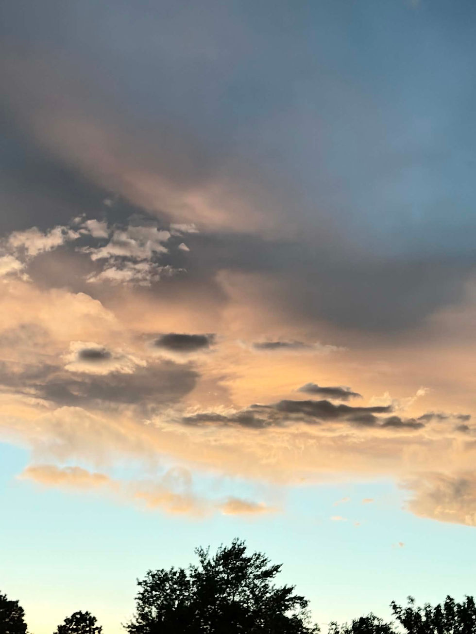 Colorful sunset sky with dramatic clouds in hues of orange and blue, silhouetted by tree tops below. Tranquil and picturesque scene.