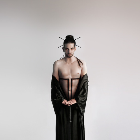 Front view of a man standing in a black silk robe that flows to the ground, partially open to reveal a bold geometric tattoo running vertically from his chest to his navel. Hair styled with paintbrushes in a topknot, evoking a modern samurai aesthetic. Tattoo by Delphine Cencig.