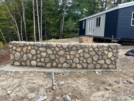 Stone wall construction with a small house in the background. TGK Hardscapes.