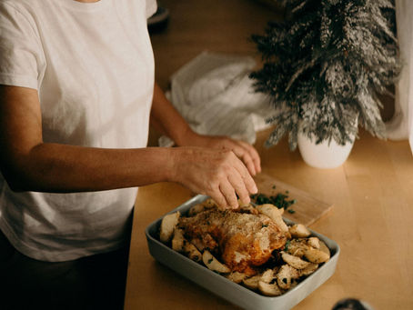 Person prepares a baked chicken with potatoes on a kitchen counter. A small, snowy tree decorates the wooden surface, creating a cozy mood.