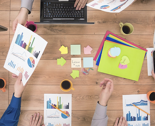 People's hands sharing and analyzing colorful charts at a wooden table with laptops, tablets, sticky notes, and coffee cups.