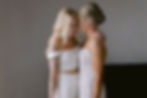 Two women in elegant white dresses laugh together, with one having tattooed arms. They stand close in a softly lit room, creating a joyful mood.