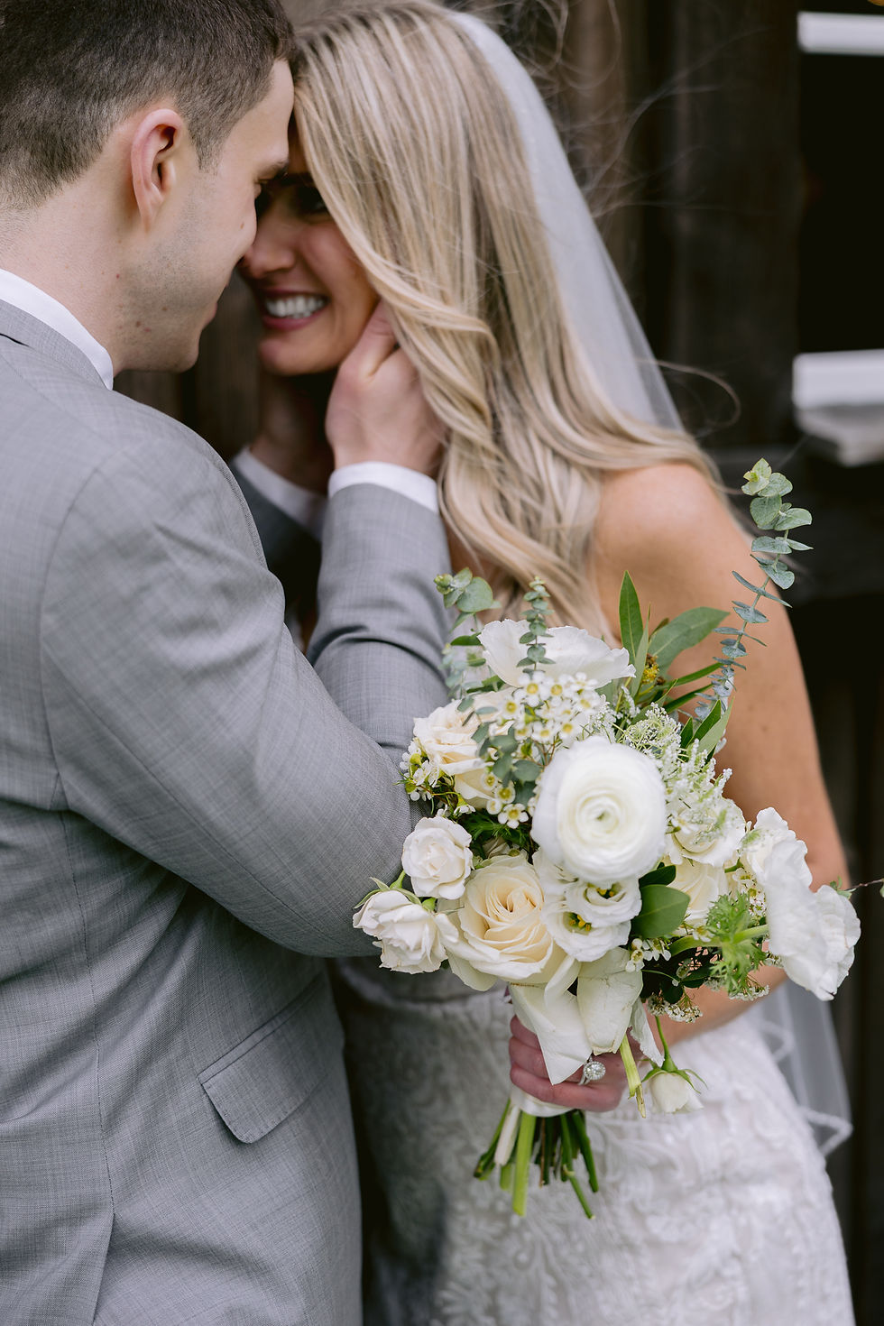 Bride and groom embrace, smiling warmly. Bride holds a white and green bouquet; groom in gray suit. Background is blurred and rustic.
