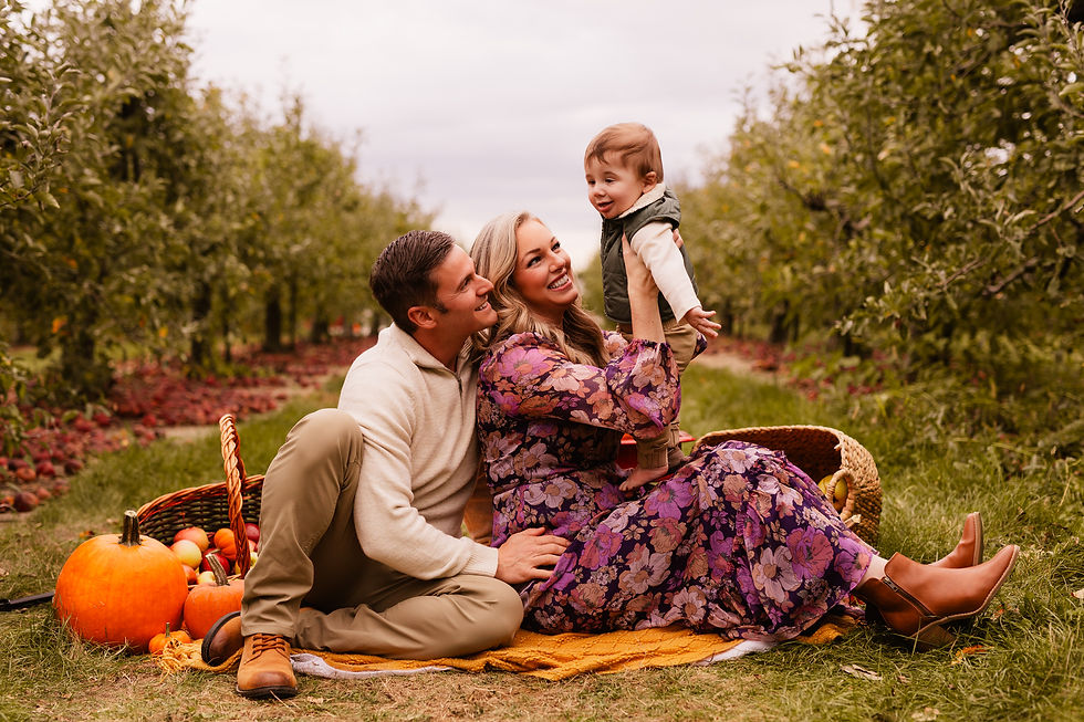 Family of three sits on a blanket in an apple orchard. Parents smile at child, surrounded by pumpkins and apples. Warm, joyful atmosphere.
