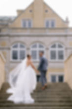 Bride in white dress and groom in gray suit holding hands, walking up steps, smiling. Background: large building with arched windows.