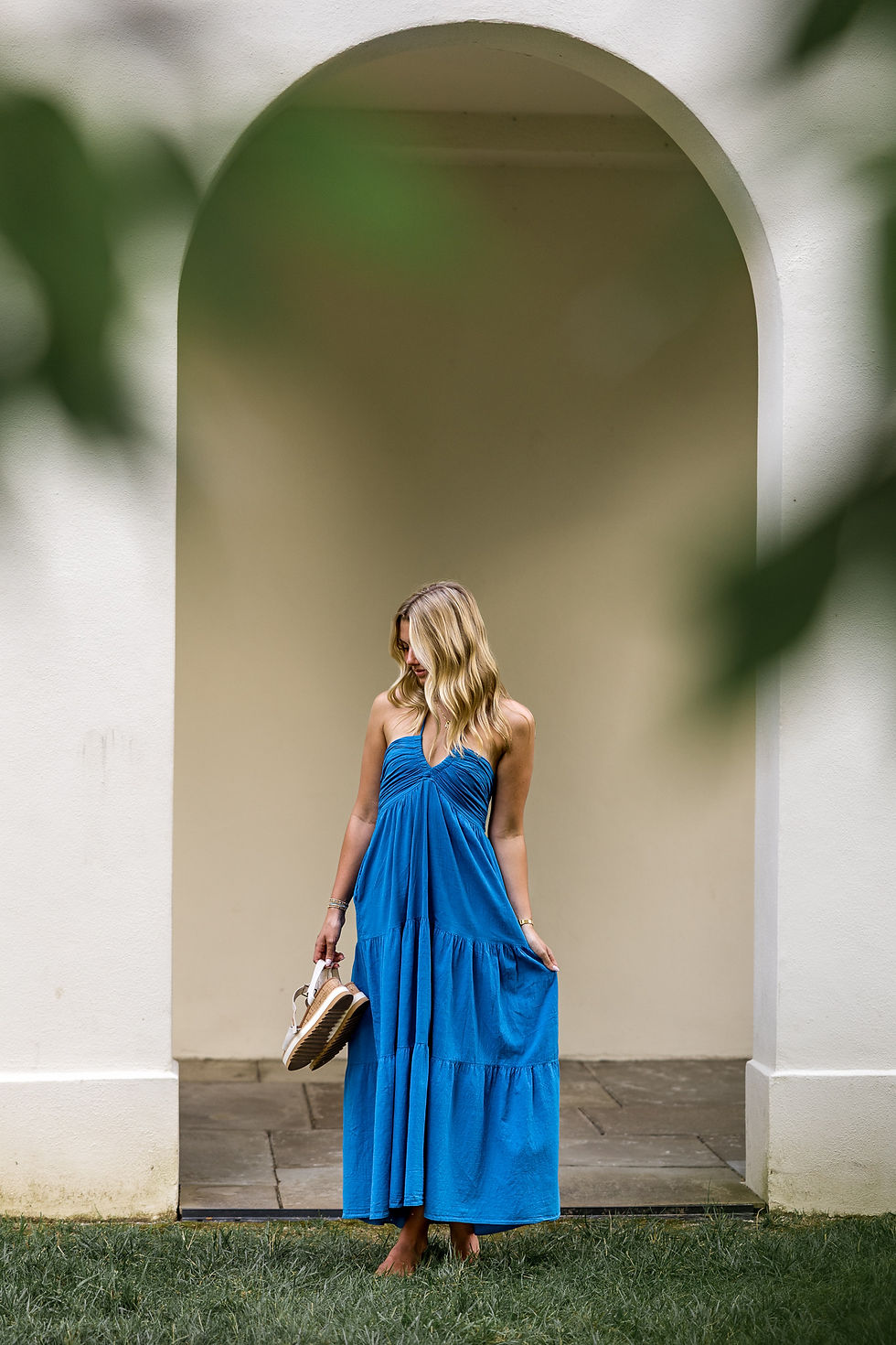 Woman in a blue dress stands barefoot under an archway, holding sandals. She gazes downward, surrounded by cream walls and green grass.