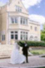 Bride and groom dance joyfully in front of a grand brick building, under a clear sky. She's in a white gown; he's in a black tuxedo.