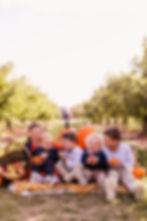 Five children sit on a blanket in an orchard with pumpkins and apples, smiling and holding small pumpkins. The setting is bright and sunny.