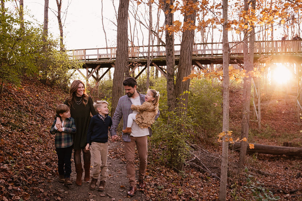 Family of five walks on a forest trail with autumn leaves, wooden bridge, and sunset in the background. Smiling, happy mood.