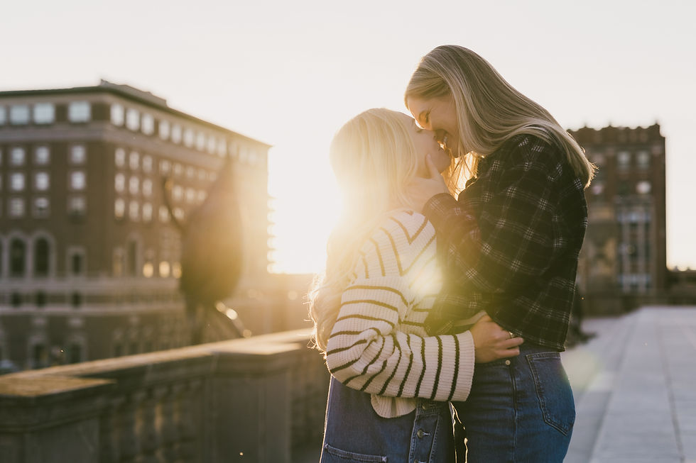 Two people embrace on a sunlit rooftop. They're smiling, with soft sunlight creating a warm glow. Urban buildings are visible in the background.