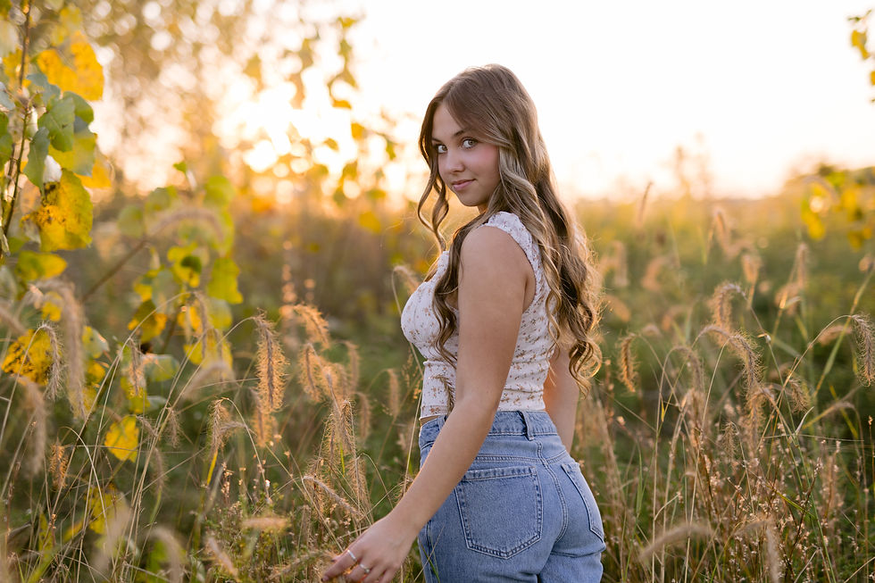 Woman with long hair in a floral top and jeans stands in a sunny meadow at sunset, surrounded by tall grass and yellow leaves.