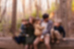 Family of five sits on a log in a forest, smiling warmly. Autumn colors fill the background, creating a cozy and joyful atmosphere.