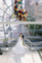 Bride and groom embrace on a spiral staircase. She wears a flowing white gown; he wears a suit. Colorful sculpture hangs above, city view outside.