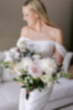Bride in an off-shoulder white dress holds a lush bouquet of pastel flowers, smiling softly. Cream couch in the blurred background.