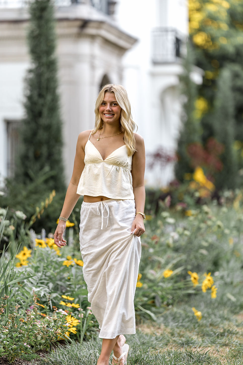 A woman in a white top and skirt walks through a garden with yellow flowers. She smiles, surrounded by greenery and a classical building.