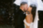 A couple kisses under a clear umbrella in the rain at night. The bride wears a white dress, and raindrops glisten against a dark backdrop.