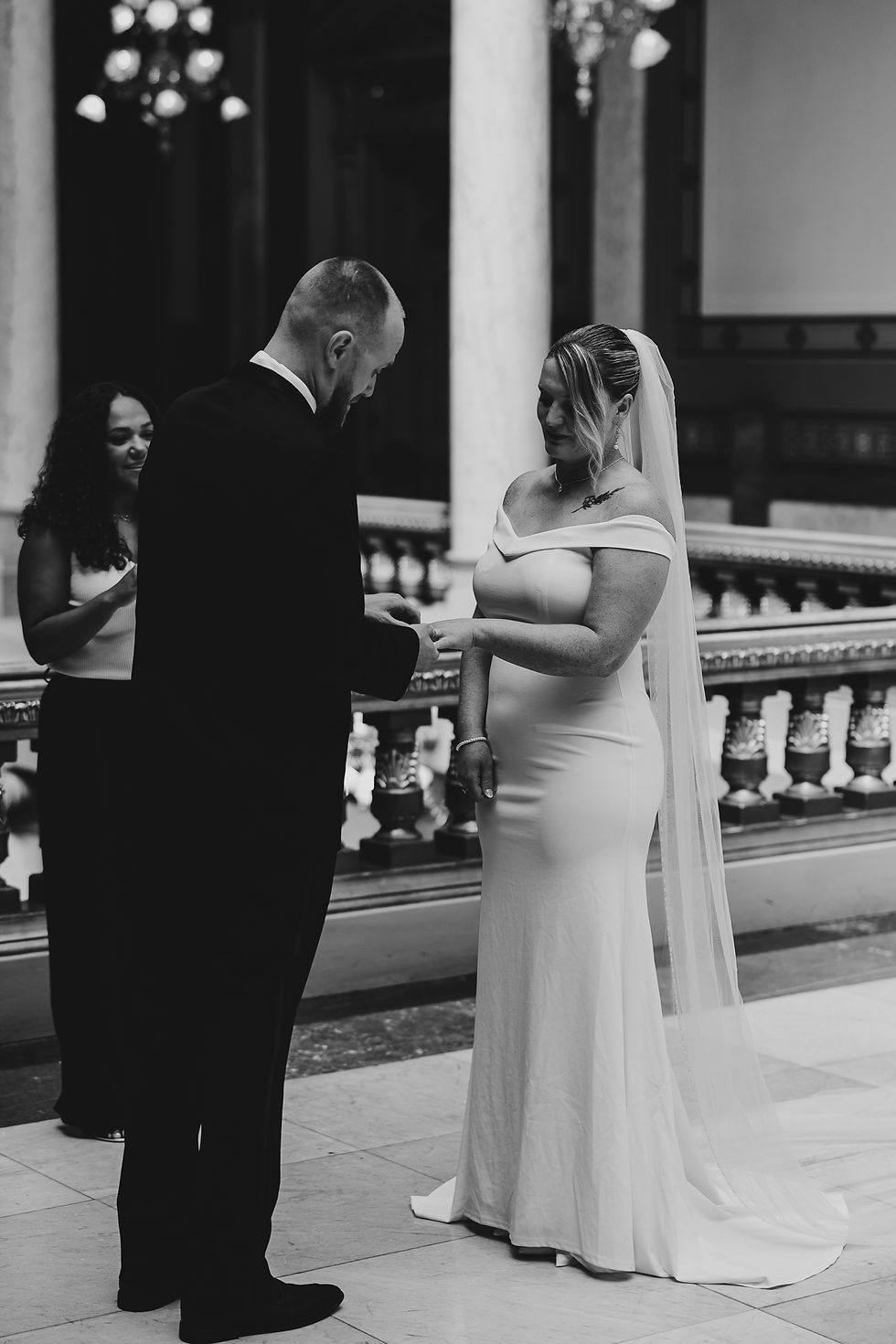 Bride and groom exchange rings in an elegant hall with chandeliers. The bride wears a veil, and an officiant stands nearby, creating a joyful scene.