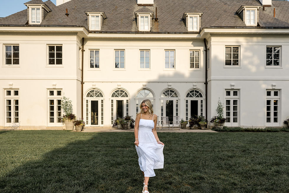 Alt text: Senior portrait of Allie in a white dress walking across the lawn in front of the historic Lilly House at Newfields in Indianapolis.