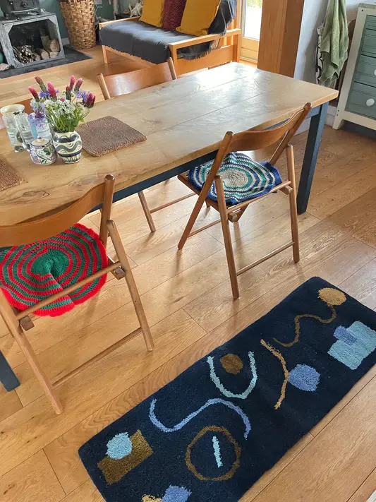 Wooden table with chairs featuring colorful cushions, vase with flowers, and blue rug with abstract patterns on wooden floor in cozy room.