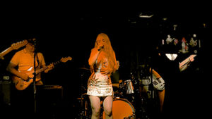 Singer with long hair performs on stage, wearing a sequin dress, holding a microphone. Drummer in background. Dimly lit, lively mood.