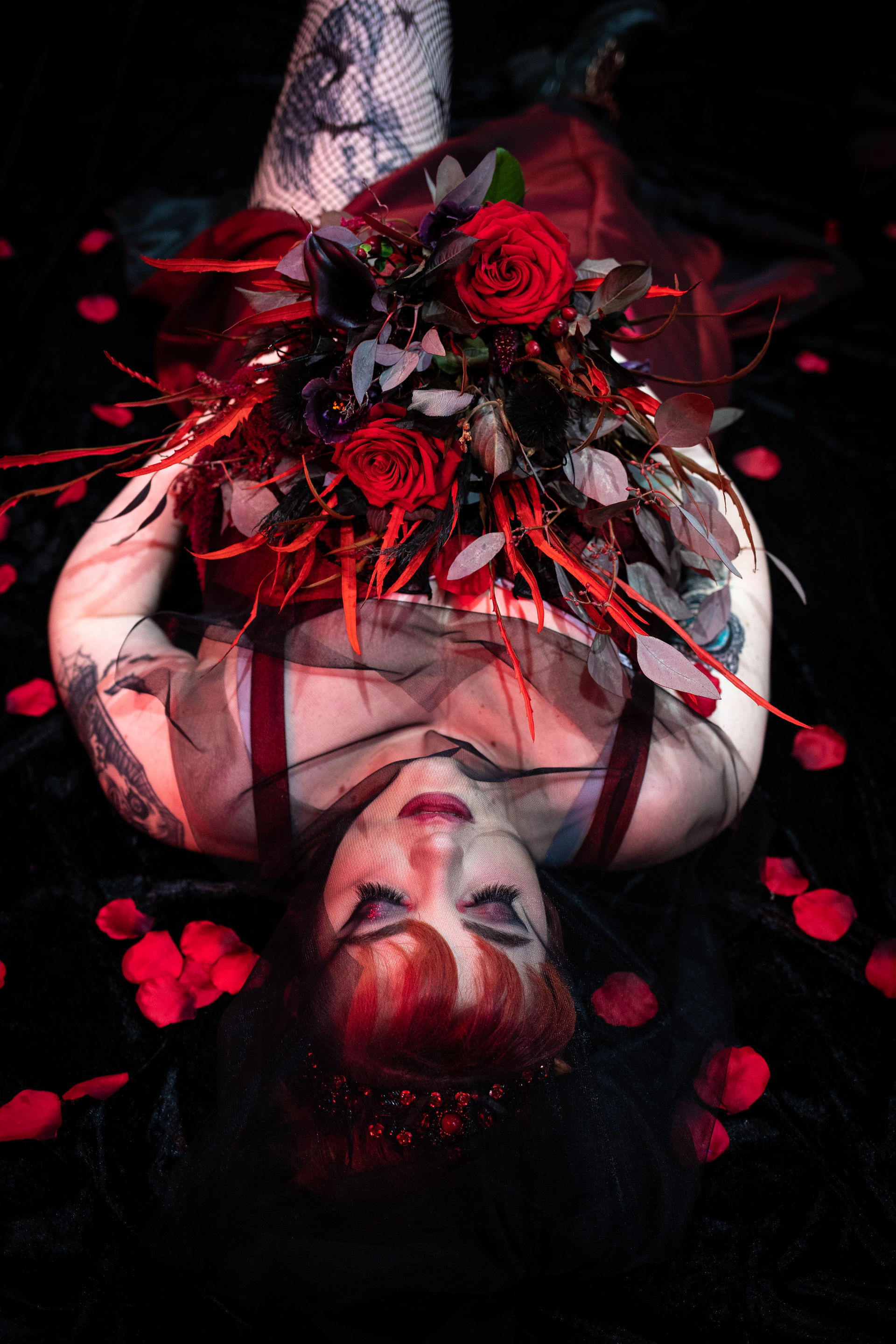 a gothic bride with bright red heair and smoky dramatic dark eye make up poses boudoir style surrounded by red rose petals