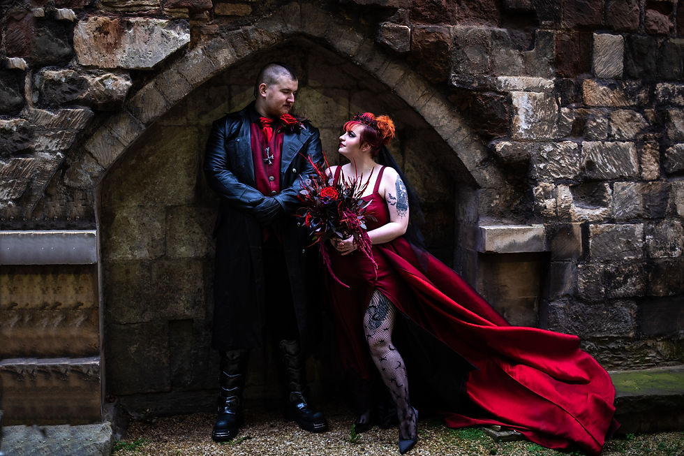 A couple dressed in gothic-style attire poses romantically by a stone arch. The woman holds a bouquet, and both wear red and black.
