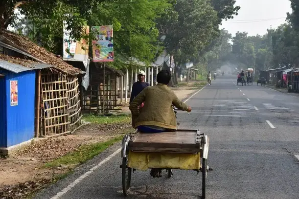 A view from a bicycle following a traditional rickshaw van down a quiet, open country highway.