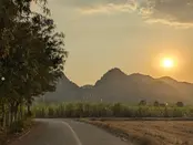 A winding country road leading toward mountain peaks during a glowing sunset in Kanchanaburi, Thailand.
